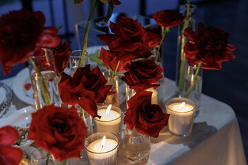 Candlelit table adorned with numerous flickering candles in glass holders and vases filled with red roses creates a warm, romantic atmosphere perfect for intimate gatherings or events.