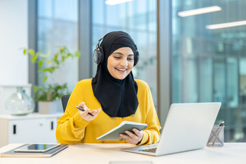 A smiling woman in a hijab, wearing headphones, attends a virtual meeting using a laptop in an...