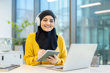 A smiling Muslim woman wearing a hijab and headphones, working on a laptop and taking notes at her...