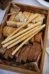 Assortment of sliced bread and breadsticks displayed in a wooden tray. Variety includes white and whole-grain bread, neatly arranged for serving or catering presentation.
