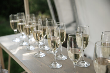 Glasses filled with sparkling wine neatly arranged on a wooden table outdoors. The setting suggests preparation for a celebration or event under natural daylight conditions.