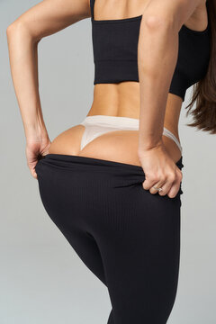 Dressed in black leggings and a matching top, a woman adjusts her waistband, revealing beige undergarments with medium skin tone. Long brown hair flows freely down her back