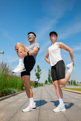Dynamic duo performs stretching exercises outdoors under a clear blue sky at a sunlit urban park in...