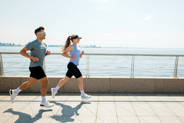 Couple jogs along a waterfront path in bright sunlight, enjoying an active and healthy lifestyle...