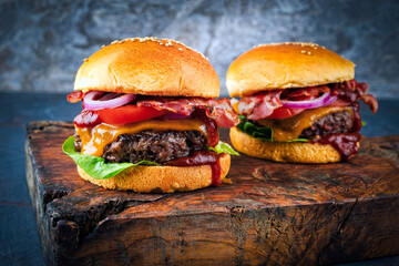 Classical cheeseburger with beef patty, cheese and bacon served as close-up with onion rings and tomato ketchup on a wooden cutting board