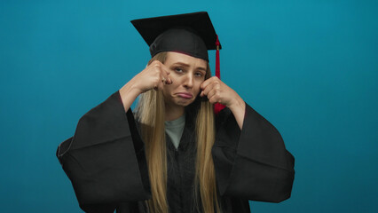 Young woman in graduation attire makes playful crying gesture against isolated blue background,...