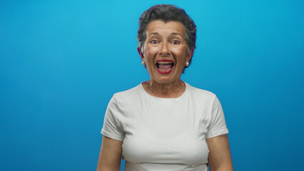 Senior woman with grey hair clapping and smiling against a vibrant blue background exuding...
