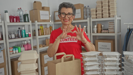 Senior woman with grey hair making heart sign in a volunteer room surrounded by donation boxes and...