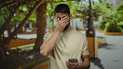 Young man in a park outdoors looking displeased at his phone, under lush green trees and sunlight.