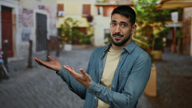 Young man outdoors shows an expression of confusion while extending hands in a charming urban street, set against a vibrant city backdrop, exuding relaxed charm in daylight.