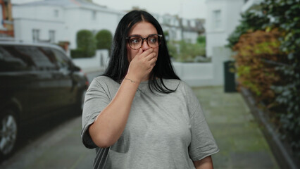 Woman with glasses stands on a city street looking surprised and curious, capturing urban outdoor...
