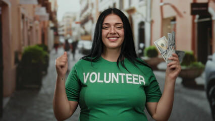 Woman smiling in city street wearing volunteer shirt holding dollars showing enthusiasm and...