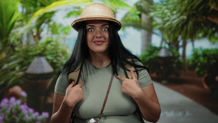 Woman tourist with surprised expression points at herself in a lush outdoor park setting, wearing a...