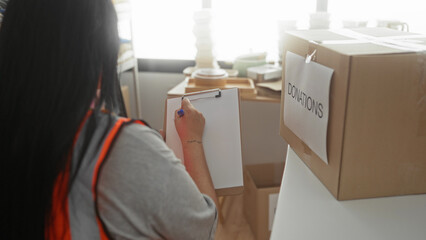 Woman writing on clipboard wearing orange vest in donation center with boxes and clothing items...