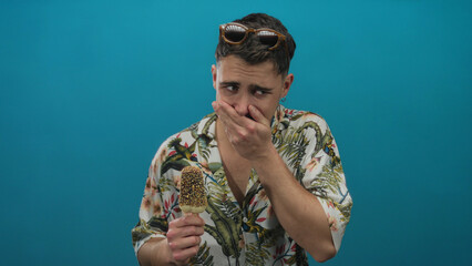 Young man holding ice cream cone in colorful shirt over blue background expressing surprise with...