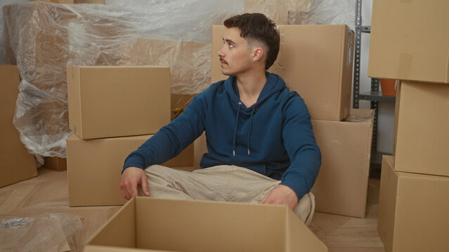 Young hispanic man sitting among cardboard boxes in a new apartment living room stretching arms in relaxed mood. - Powered by Adobe