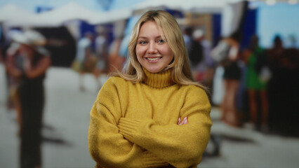 Smiling woman with long blonde hair and yellow sweater stands with arms crossed in a lively outdoor...