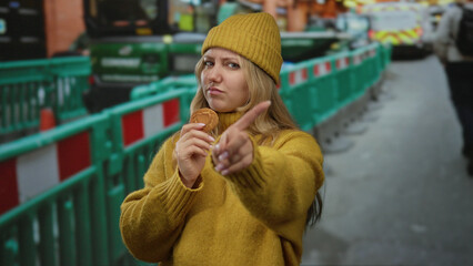 Woman in yellow sweater and beanie points finger at construction site, holding cookie, with...
