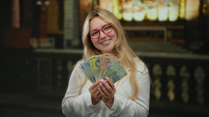 Woman holding romanian banknotes smiling inside church environment with money and colorful stained glass in background enhancing spiritual wealth contrast.