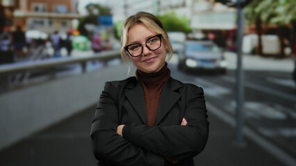 Confident young woman with blonde hair and glasses stands on a bustling city street terrace, arms...