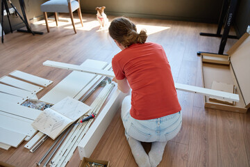 Woman assembling white furniture parts on wooden floor using drill, work tools and instruction...