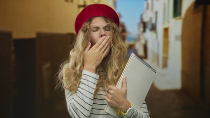Woman covering hand while yawning on street in blonde young outfit holding notebook wearing glasses...