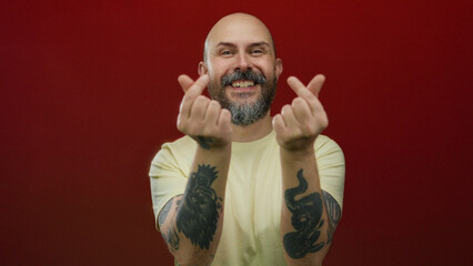 Bald, tattooed man with a beard making a heart gesture against a bright red wall, showcasing a casual, friendly expression.