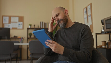 Man with beard using tablet in modern office interior, looking thoughtful, wearing casual sweater,...