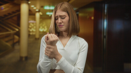 Young blonde woman grips her bare wrist in pain gesture inside hotel corridor; discomfort injury...