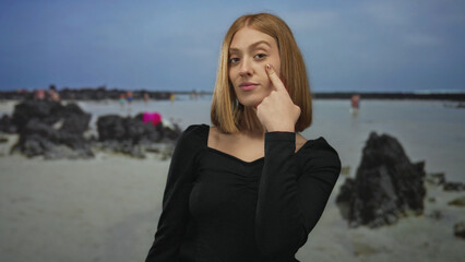 Young blonde woman pointing finger to eye at a sunny seaside beach with clear horizon; observation...