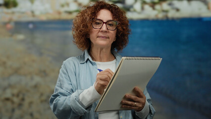 Fototapeta premium Woman writing in notebook on seaside capturing the serene beach atmosphere while the ocean forms a picturesque backdrop under the clear blue sky.