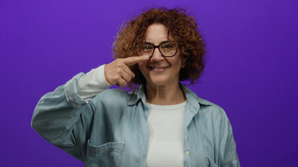 Hispanic woman with curly brunette hair and glasses pointing at her nose with a playful smile in front of a vibrant purple isolated background.