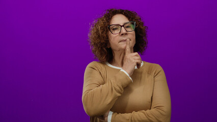 Woman pondering with hand on chin against vibrant purple background, wearing brown sweater, curly...