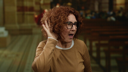 Woman with curly hair and glasses expressing surprise inside a dimly lit church wearing a brown...