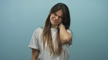 Hispanic woman in white t-shirt expresses neck pain over isolated blue background wall, capturing a...