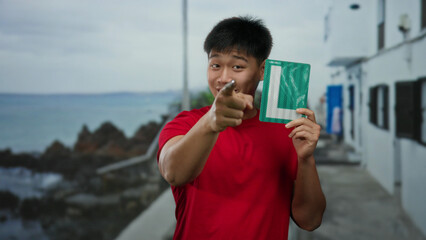 Young man on seaside promenade holding learner sign outdoors with a confident smile and index finger pointing towards the camera against a scenic coastal backdrop.