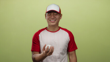 Young man in red baseball uniform and cap smiling over yellow background, holding glove and ball,...
