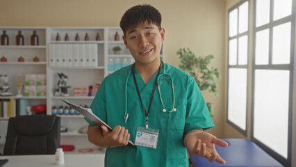 Young asian male doctor in green scrubs holding a clipboard in a hospital clinic room suggesting a...