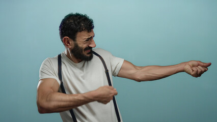 Hispanic man with beard pretending to pull rope against plain blue background, showcasing humorous...