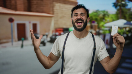 Hispanic man with beard joyfully holds credit card on bustling street outdoors, embodying financial...