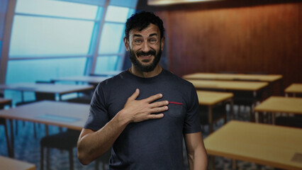Hispanic man with beard expressing gratitude by touching his chest, standing in a school room with tables in the background.