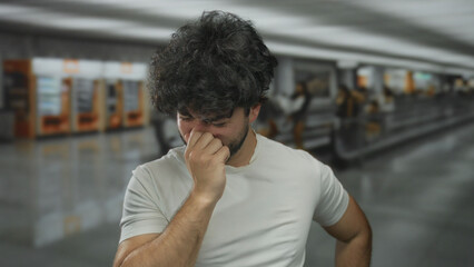 Young hispanic man with beard sneezing indoors in airport setting, capturing a candid moment of discomfort in an airport terminal environment.