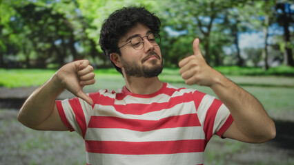 Young man with beard in striped shirt making thumbs up and down gestures in a green park outdoors...