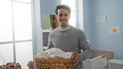 Young man smiling while holding laundry basket indoors in bright room with books and toys visible in background suggesting a home or apartment setting.