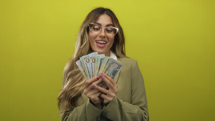 Businesswoman holds fan of dollar bills in both hands at her face in a studio with yellow backdrop;...