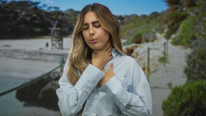 Blonde woman clutch throat and chest with closed eyes on sunny sandy beach walkway; panic distress...