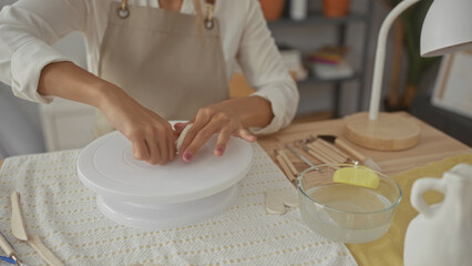 Woman kneads clay with her bare hands on a spinning wheel in a pottery studio while wearing a beige...