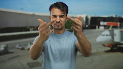 Young hispanic man making rude gestures at an airport with blurred airplanes in the background,...