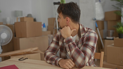 Young man in new apartment surrounded by boxes, wearing plaid shirt, sitting at table,...