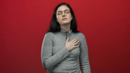 Woman in glasses and gray sweater standing against vibrant red background, looking reflective with...
